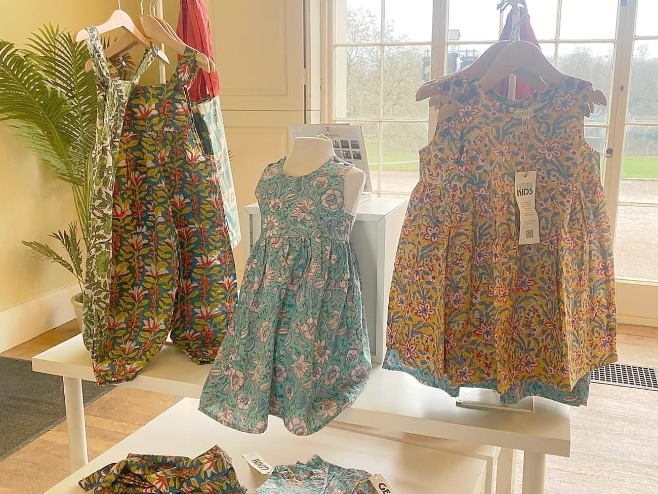 Genki Kids dresses and dungarees on a display table in the English Heritage shop at Kenwood House, with the sash windows and parkland behind